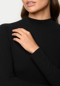 Schwarzes langärmliges Oberteil mit hohem Kragen, glatter Textur und taillierter Passform. Eine Hand ruht sanft auf dem Schulterbereich.