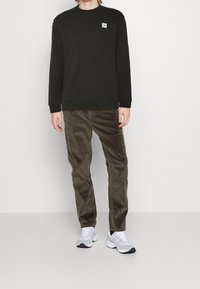 Sudadera negra con cuello redondo, combinada con pantalones de pana verde oliva. Presenta un pequeño parche de logo azul. Calzado: zapatillas blancas y grises.