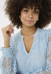 Světle modrá krajková blůza s výstřihem do V a nařasenými bufantovými rukávy, která má složité květinové vzory a průsvitnou texturu.