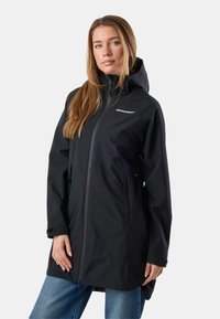 Žena v čiernej pršiplášti s kapucňou, so zipsom vpredu a vreckami, stojaca na jednoduchom svetlom pozadí.