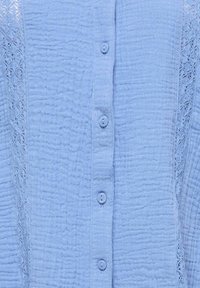 Tissu bleu clair avec fermeture verticale à boutons, motif froissé texturé, et inserts en dentelle de chaque côté.