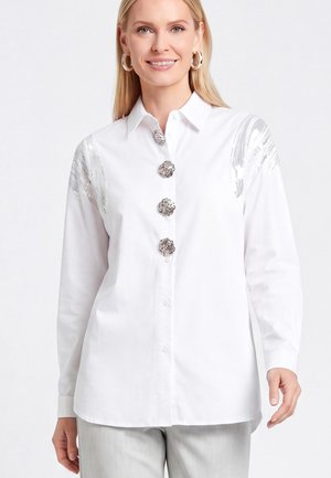 Donna sorridente che indossa una camicetta bianca con grandi bottoni decorativi argento e dettagli in paillettes argento sulle spalle, abbinata a pantaloni grigio chiaro.