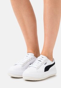 White sneakers with a textured sole, featuring black suede accents and a padded tongue. Laces in white, with perforations on the sides.