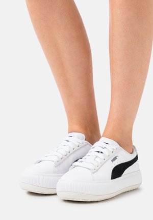 White sneakers with a textured sole, featuring black suede accents and a padded tongue. Laces in white, with perforations on the sides.