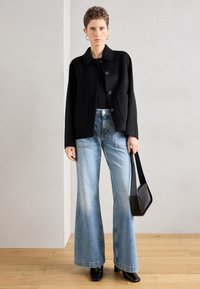Casaco preto de mistura de lã com gola e botões, combinado com jeans azuis claros de corte flare. Modelo segura uma mala preta e usa sapatos pretos com salto.