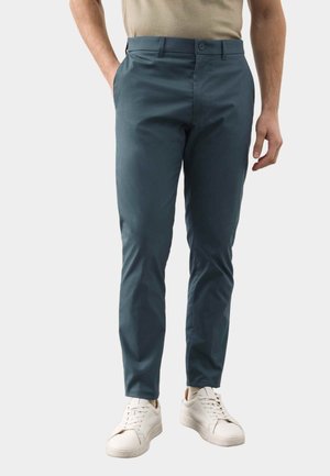 Hombre con pantalones ajustados azul oscuro, camisa beige y zapatillas blancas, de pie con una mano en el bolsillo sobre fondo blanco.