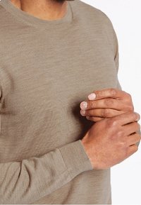 Khaki svetřík s dlouhým rukávem a kulatým výstřihem, vyrobený z jemné pleteniny. Má žebrované manžety a hladkou strukturu. Detail rukou držících látku.