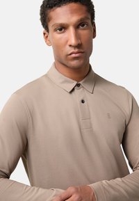 Beige polo tričko s dlouhým rukávem a límečkem, s poklopcem na tři knoflíky a jemně vyšitým logem na hrudi. Hladká textura.