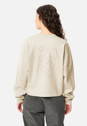 Frau mit lockigem Haar steht mit dem Rücken zur Kamera und trägt einen hellbeigen Pullover mit abstrakter Linienkunst und dem Text "outdoor 24/7" auf dem Rücken.