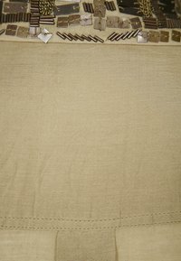 Tejido beige con acentos metálicos, incluyendo formas cuadradas y rectangulares, y detalles texturizados cerca de la parte superior, con costuras a lo largo de la parte inferior.
