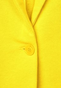 Žlutý pletený kardigan s výrazným žlutým knoflíkem, hladkou strukturou a výstřihem do V.