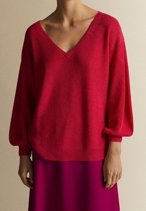 Kobieta ubrana w luźny czerwony sweter z dekoltem w serek i bufiastymi rękawami oraz głęboko różową spódnicę na jednolitym beżowym tle.