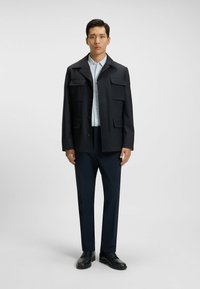 Chaqueta ligera de la marina con múltiples bolsillos, superpuesta a una camisa de botones azul y combinada con pantalones oscuros y zapatos negros.