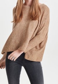 Světle hnědý oversized pletený svetr s žebrovanými manžetami, širokým výstřihem a uvolněnou siluetou, spárovaný s tmavými slim džínami.