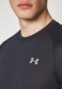 Detailní záběr na osobu v černém tričku Under Armour s světle šedým logem na hrudi.