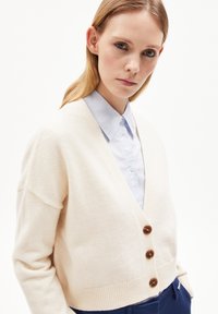 Crème cardigan met een V-halsontwerp, voorzien van drie bruine knopen, geribbelde zoom en losse mouwen, gedragen over een lichtblauw overhemd met kraag.
