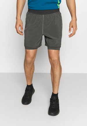 Homme portant un short de sport gris, des baskets noires et une chemise bleue, debout les mains relâchées le long du corps sur un sol uni.