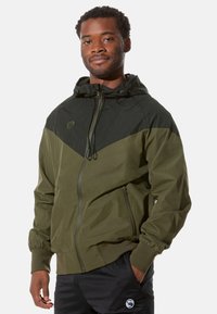 Olivgrüne Jacke mit schwarzem Brustbereich, Frontreißverschluss, verstellbarem Kapuzen und elastischen Bündchen, mit einem Logo auf der Brust.