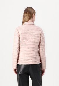 Ροζ καπιτονέ μπουφάν με όρθιο γιακά, που διαθέτει οριζόντια ραφή και ομαλή υφή. Φορεμένο πάνω από μαύρο παντελόνι.