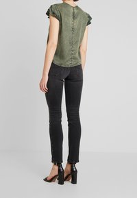Mujer vestida con una blusa de manga corta verde oliva con bordes fruncidos, jeans skinny negros y sandalias de tacón negro, de pie contra un fondo gris.