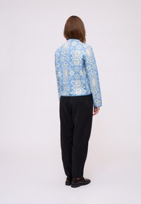 Persona con lunghi capelli castani in piedi di spalle, indossa una giacca blu a fantasia, pantaloni neri e scarpe marroni scure su sfondo plain.