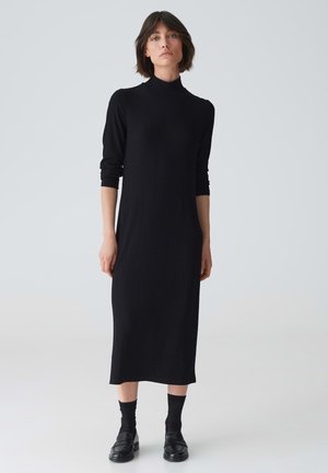 Robe midi noire à côtes avec un col haut et des manches longues, présentant une silhouette ajustée et une texture douce. Associée à des chaussettes noires et des mocassins.