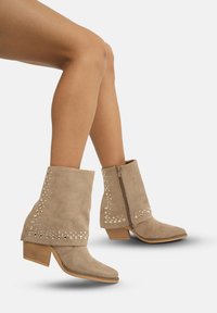 Botas de tobillo de ante beige con punta afilada, adornos decorativos con tachuelas y cremallera lateral, que cuentan con un tacón grueso y una textura suave.