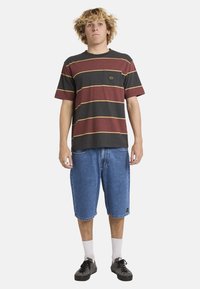 Junger Mann mit lockigem blondem Haar, der ein bordeauxrotes und schwarz gestreiftes T-Shirt, lange Jeansshorts, weiße Socken und schwarze Turnschuhe trägt.