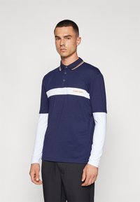 Ναυτικό polo μπλουζάκι με κοντά μανίκια, λευκή ρίγα διαγώνια στο στήθος, πορτοκαλί λογότυπο και αντίθετο γιακά. Μοντέλο το φοράει με σκούρα παντελόνια.