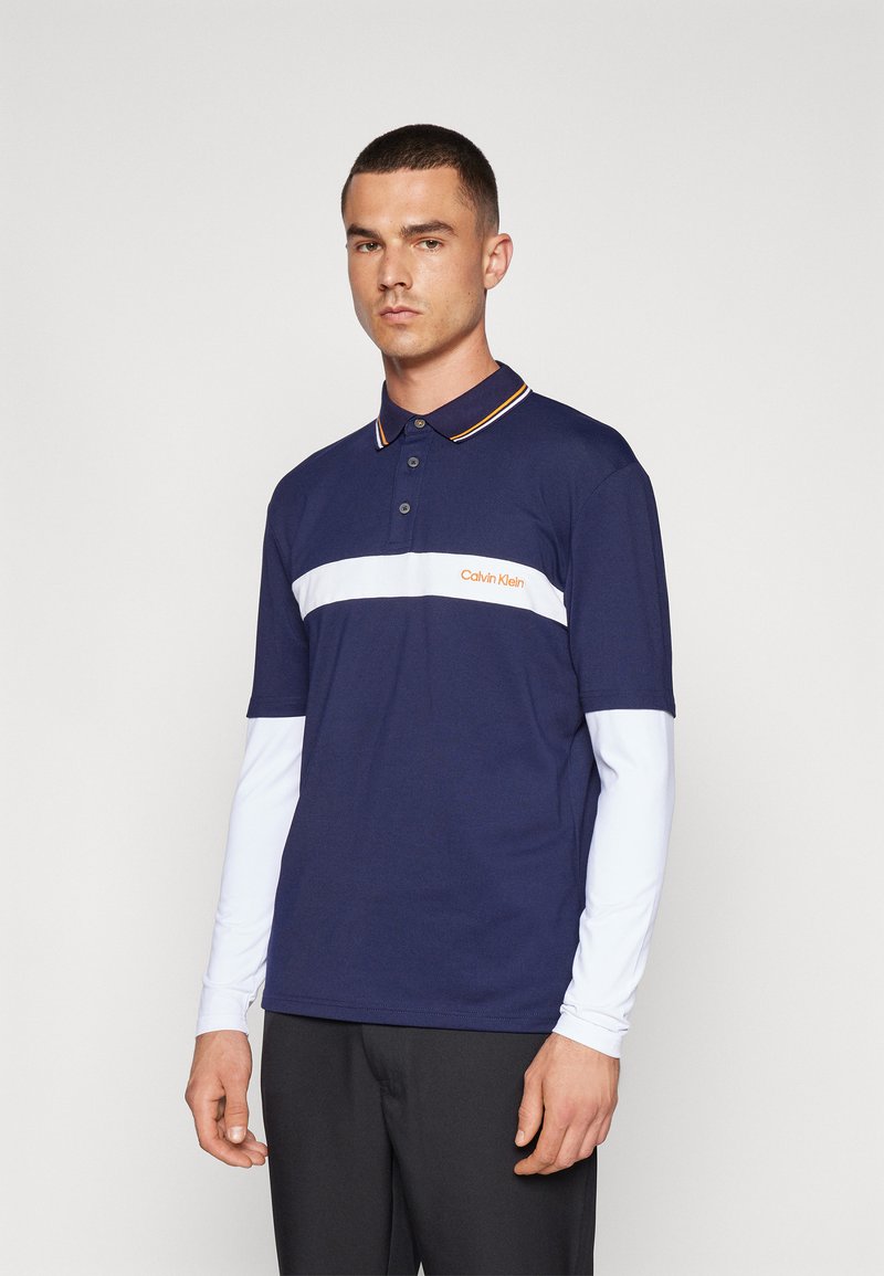 Ναυτικό polo μπλουζάκι με κοντά μανίκια, λευκή ρίγα διαγώνια στο στήθος, πορτοκαλί λογότυπο και αντίθετο γιακά. Μοντέλο το φοράει με σκούρα παντελόνια.