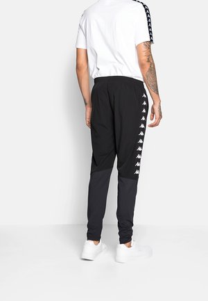 Mand iført sorte tapered træningsbukser med hvide logoer ned langs siderne, hvide sneakers og en hvid t-shirt med logostiber på ærmerne, stående.