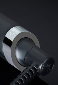 Outil de coiffure noir avec un col en chrome métallique et un manche noir texturé. Il présente le logo en relief "ghd" avec des accents argentés.
