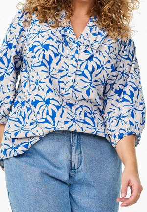 Woman wearing a blue and white patterned blouse with ruffled collar and blue jeans, standing with curly hair partially visible.