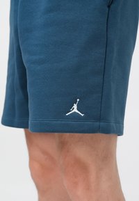 Shorts de sport bleu foncé en tissu doux, avec un logo Jordan blanc brodé sur l'ourlet inférieur. Texture lisse et design décontracté.