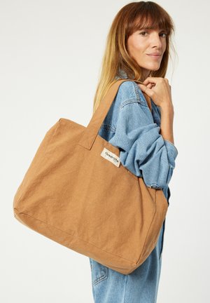 Große braune Canvas-Tasche in rechteckiger Form, mit robusten Tragegriffen und einem seitlichen Etikett mit dem Markennamen. Weiche Textur, keine sichtbaren Muster.