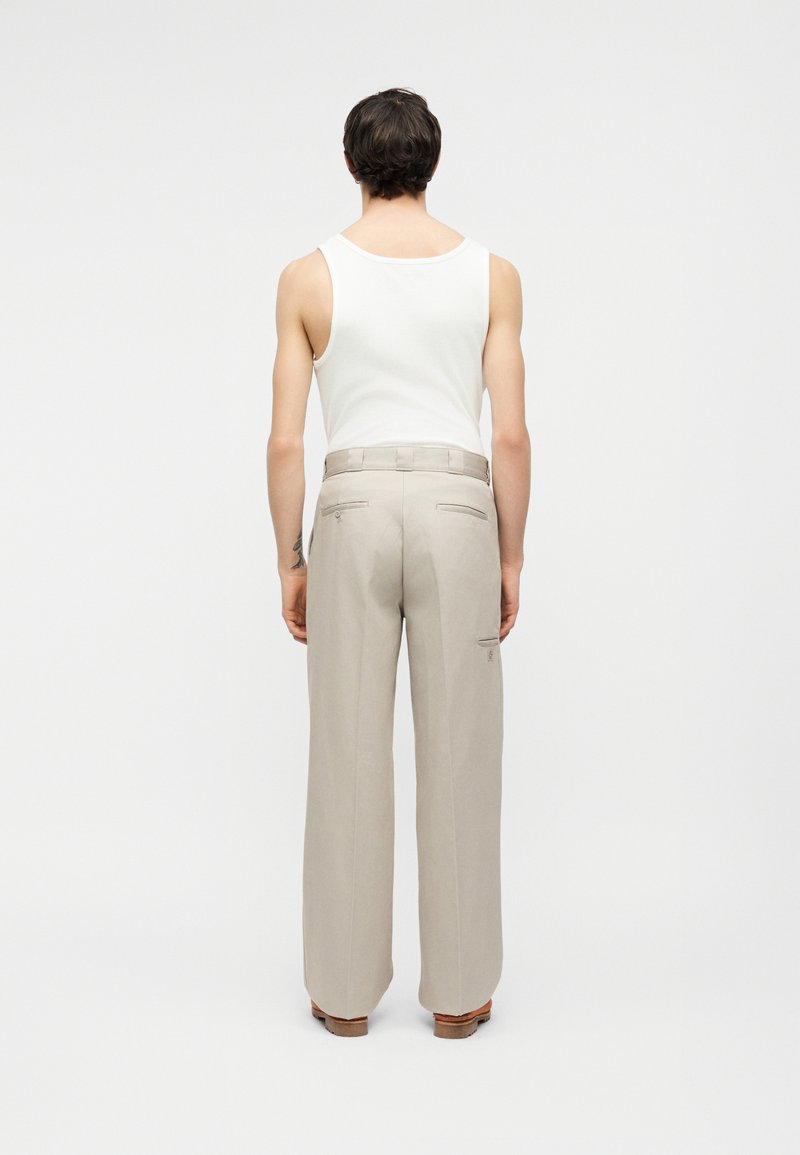 Personne aux cheveux courts et foncés portant un débardeur blanc et un pantalon large beige, debout de dos sur un fond uni.