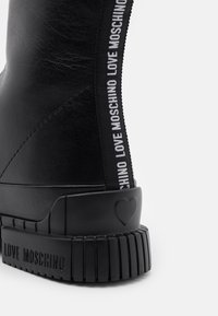 Černé kotníkové boty s hladkým koženým povrchem, vybavené bílým elastickým bočním panelem s logem a robustní černou gumovou podešví s detailem srdce.