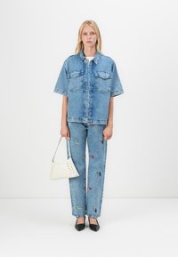 Jachetă din denim cu mâneci scurte și două buzunare pe piept, asociată cu blugi brodați de culoare albastră cu modele colorate și o geantă albă.