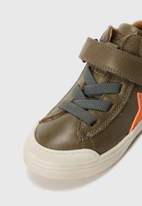 Grønn lærersneaker med grå snøring, en borrelåsrem og et oransje accent på siden. Gummisåle med teksturdetaljer.