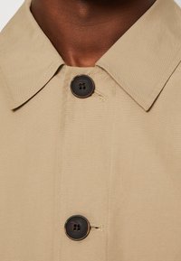 Khaki jas met een gestructureerde kraag, voorzien van een donkere knop en zichtbare stiksels op een gladde, lichte stof.