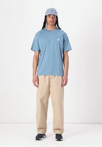T-shirt bleu clair à manches courtes avec un logo, pantalon beige ample, et une casquette bleu clair. Porté avec des chaussures de sport noires.