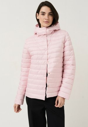 Femme aux cheveux foncés portant une veste matelassée rose clair avec capuche, debout devant un fond clair uni, mains détendues le long du corps.