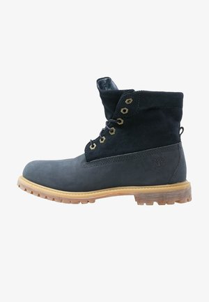 Botte à lacets en cuir noir et suède avec semelle en caoutchouc beige et œillets en métal, logo Timberland sur le talon, vue de côté.