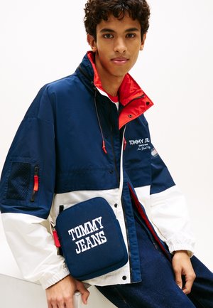 Joven con una chaqueta azul marino, blanca y roja y un bolso bandolera azul marino a juego con la marca "Tommy Jeans".