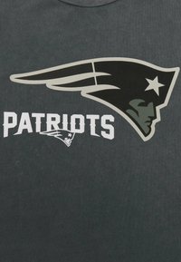 Tmavě šedé tričko s velkým černým logem klobouku, se hvězdou a slovem "PATRIOTS" v tučném bílém písmu pod ním.