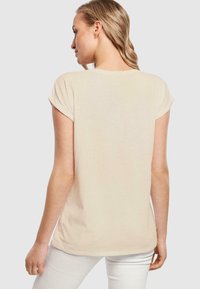 Beige t-skjorte med korte ermer laget av myk stoff. Har rullet ermer og en buet kant bak, vist fra bakvinkelen.