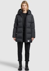 Jeune femme portant un long manteau en duvet noir avec une capuche, un pantalon noir et des bottines noires, se tenant contre un fond clair.