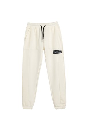 Pantaloni jogger crema realizzati in tessuto morbido, con vita elastica e coulisse, due ampie tasche laterali e un'etichetta nera decorativa.