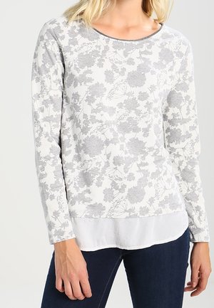 Mujer con una blusa blanca de manga larga con estampado floral gris claro, superpuesta sobre una camisa blanca, combinada con jeans azul oscuro.