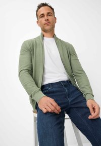 Light green zip-up cardigan made of textured knit fabric, worn over a white T-shirt. Paired with dark blue jeans, seated on a stool.